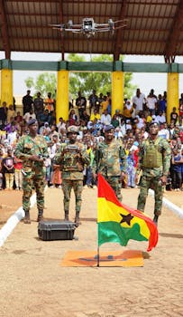 Four Ghanaian soldiers in uniform operate a drone at an outdoor public event with a Ghana flag.