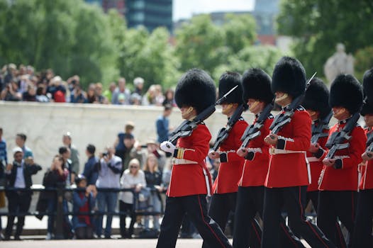 Royal guards in ceremonial red uniforms seen marching with rifles, with spectators in the background.