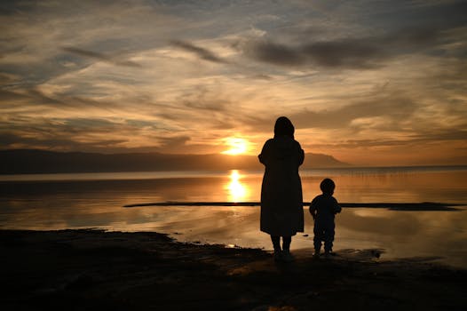 A woman and child watch a stunning sunset, creating a serene silhouette by the sea.
