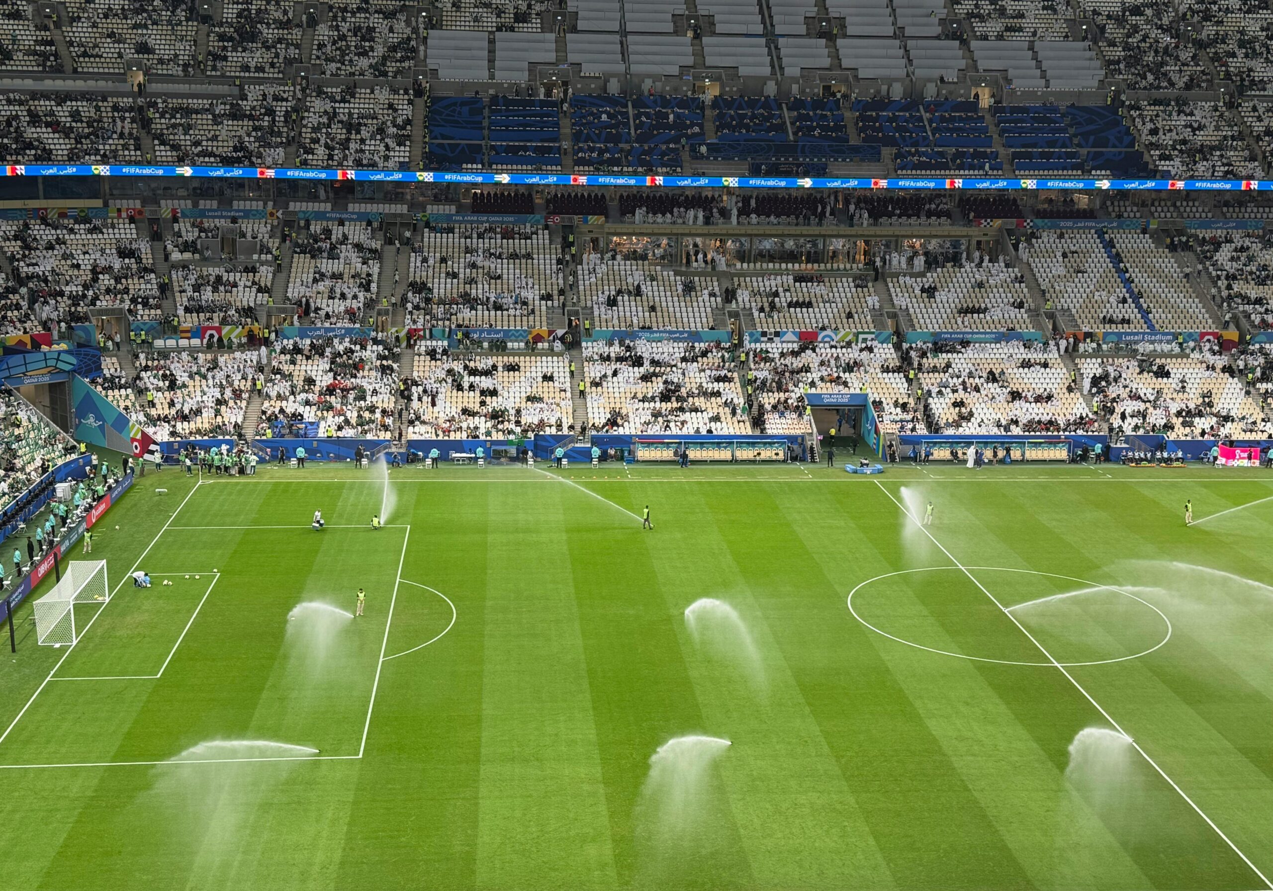 Soccer stadium field being watered with sprinklers under cloudy skies during daytime.