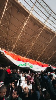 A vibrant crowd of soccer fans at Basra stadium cheering with an Iraqi flag.