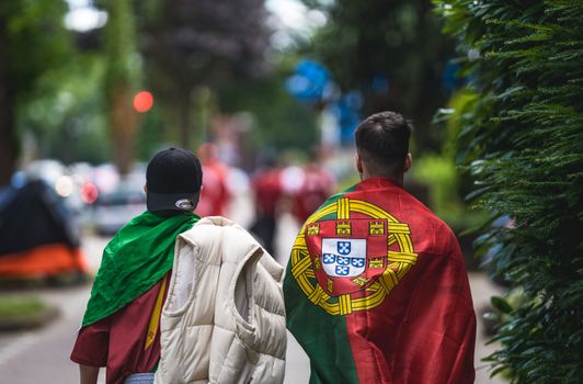 Two sports fans draped in Portuguese flags walking on a city street during daytime.