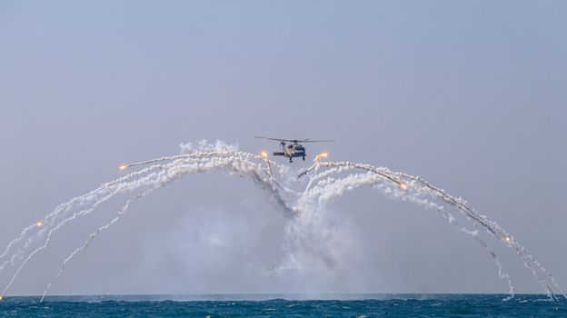 A helicopter executes a stunning aerial maneuver, releasing flares over the sea against a clear sky.