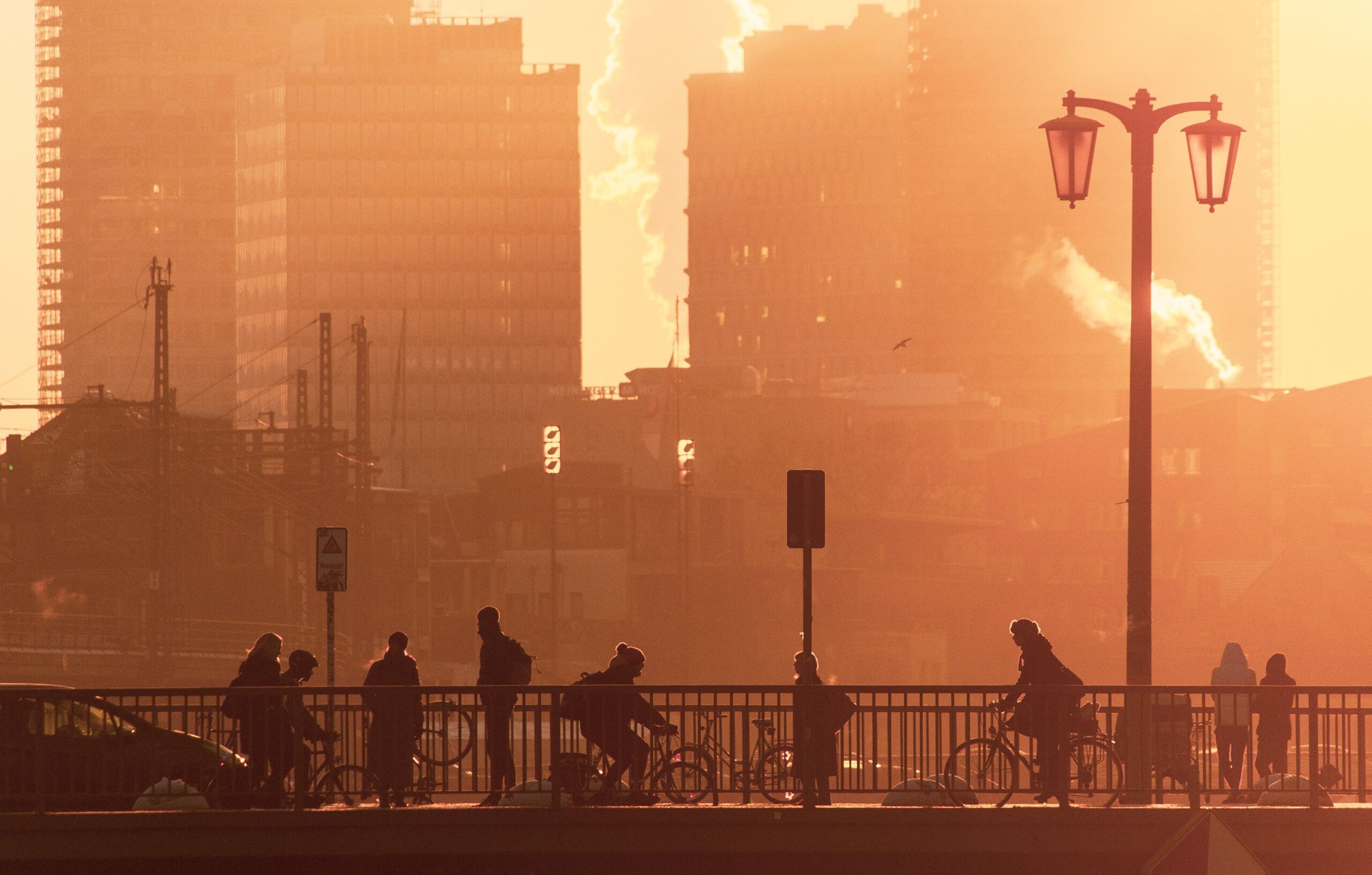 Silhouetted cyclists and pedestrians cross a city bridge at sunrise, with a skyline backdrop.