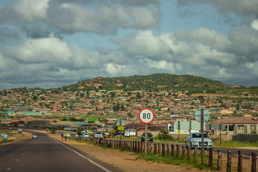 A vibrant countryside town nestled by mountains and a scenic road with vehicles under a blue sky.