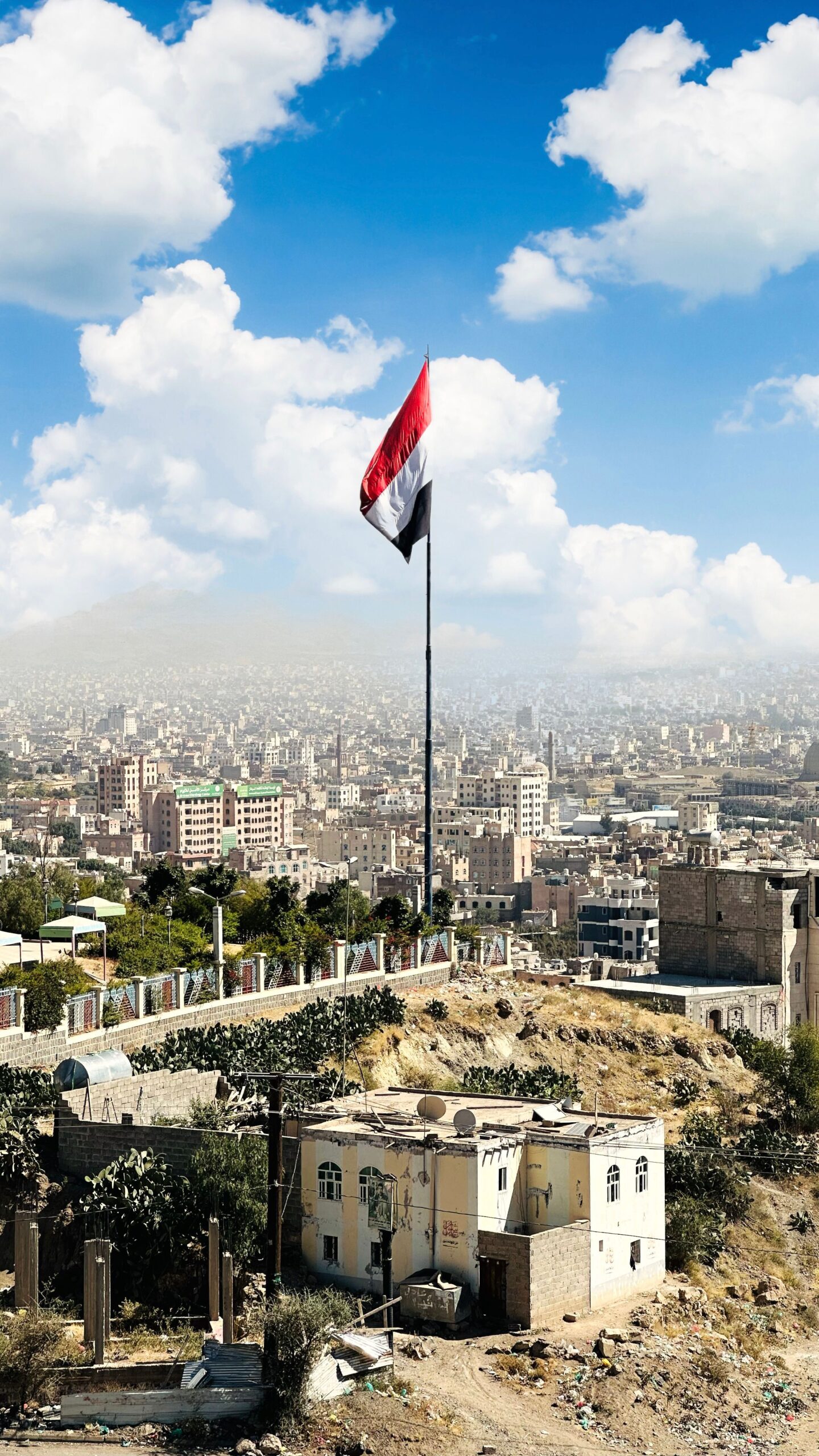 Elevated view of Sana'a cityscape with prominent Yemeni flag under a bright blue sky.